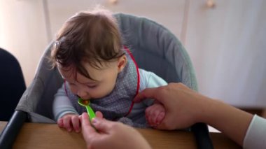 Anne isteksiz bebek besliyor, hasta katı yiyecekler sunuyor, bebek aşağı bakıyor, kaşıkla ilk deneyim, hassas bakım, evde aile büyütme anı.