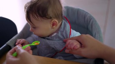 Anne bebeğe nazikçe yemek veriyor, bebeğin annesinin elini tutarken yakın plan, isteksiz yüz ifadesi, ilk kez katı gıda deneyimi, sabır ve bakım, besleyici çevre.