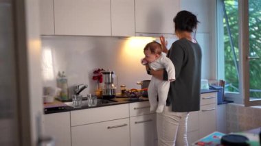 Anne mutfakta yemek hazırlarken bebeğini kucağında tutuyor, ocakta yemek pişiriyor, ev sorumluluklarını dengeliyor ve evde bakım yapıyor.