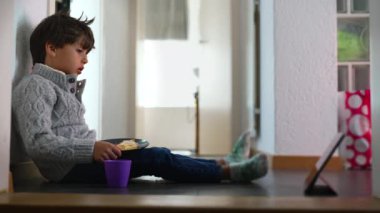 Genç çocuk duvara yaslanmış, ekmek yiyor ve içiyor, tablet izliyor, sakin ve odaklanmış, evde yemek yiyor, çocuk atıştırma zamanı, gündelik ve rahat, ev ortamı
