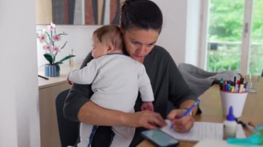 Anne, akıllı telefon kullanarak bebeğini kucağında tutuyor ve masa başında notlar alıyor. Bakıcılık görevini tamamlamaya odaklanırken aynı zamanda ev idaresini de dengeliyor.