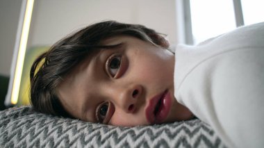 Kanepede dinlenen genç bir çocuk, sakin, yansıtıcı bir iç mekanda bir çizgi filmi kapalı izlerken yüzünün yakın çekimi.