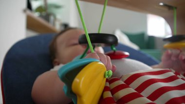 Bebek uzanıp renkli asılı oyuncaklarla oynarken, bir fedainin içinde uzanırken, duyusal aktiviteleri gösteriyor, nesneleri keşfediyor ve eğlenceli bir şekilde etkileşime geçiyor.