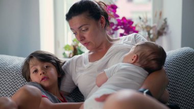 Anne, bebeği ve oğluyla kanepede dinleniyor sevgi dolu jestler ve rahat bir yuva ortamında ısınma duygusuyla aile bağlarını güçlendiriyor.