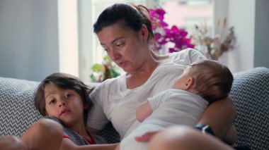 Anne, bebeği ve oğluyla kanepede dinleniyor sevgi dolu jestler ve rahat bir yuva ortamında ısınma duygusuyla aile bağlarını güçlendiriyor.