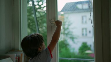 Pencereyi açmaya çalışan ama açamayan genç bir çocuk, merak ve çaba anı, evin penceresinden dış dünyaya bakan bir manzara ile iç mekan sahnesi.