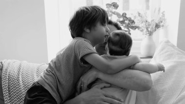 Küçük bir çocuk, kanepede hassas bir aile anı sırasında annesinin kollarında sevgi dolu bir şekilde kardeşini kucaklıyor. Güçlü bir kardeş bağı ve şefkat sergiliyor.