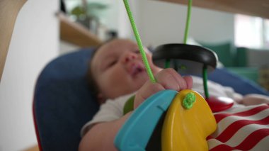 Bebek uzanıp renkli asılı oyuncaklarla oynarken, bir fedainin içinde uzanırken, duyusal aktiviteleri gösteriyor, nesneleri keşfediyor ve eğlenceli bir şekilde etkileşime geçiyor.