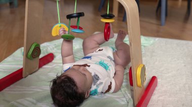 Bebek bir oyun matı üzerinde uzanıyor ve renkli asılı oyuncaklara uzanıyor, sıcak bir aile ortamında oyun saatlerinde erken gelişim ve duyusal keşif ile meşgul oluyor.