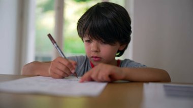 Çocuk kalemle dikkatlice çizim yapıyor, masaya yaslanıyor, dikkatle kağıda çiziyor, sanat eserlerini yaratmaya odaklanıyor.