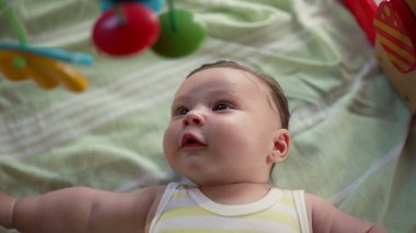 Bebek bir oyun hasırının üzerinde yatıyor, gözleri faltaşı gibi açık, renkli asılı oyuncakları inceliyor, oyuncu ve duyusal gelişimle meşgul, bir ev ortamında erken çocukluk keşfi.