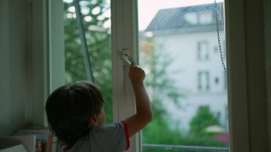 Pencereyi açmaya çalışan ama açamayan genç bir çocuk, merak ve çaba anı, evin penceresinden dış dünyaya bakan bir manzara ile iç mekan sahnesi.