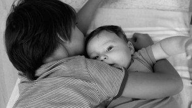 Küçük bir çocuk kardeşiyle kucaklaşıyor, rahat ve neşeli bir anda yan yana uzanıyor, evdeki hassas bir aile etkileşimini yakalıyor, siyah ve beyaz.