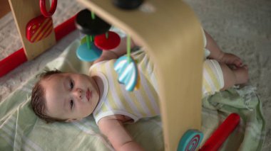Bebek bir oyun hasırının üzerinde uzanıyor, renkli asılı oyuncakları gözlemliyor, şaşkın ve meraklı, sakin bir ev ortamında erken duyusal keşifler ve oyun zamanı.