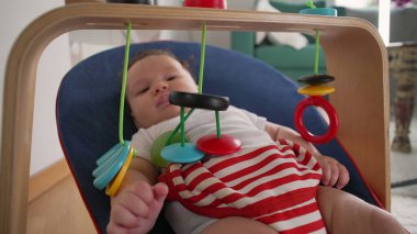Bebek bir fedaide uzanıyor, renkli asılı oyuncaklarla gülümsüyor ve etkileşimde bulunuyor, merak ve bağlılık gösteriyor, rahat bir ev ortamında, duyusal oyun odaklı.