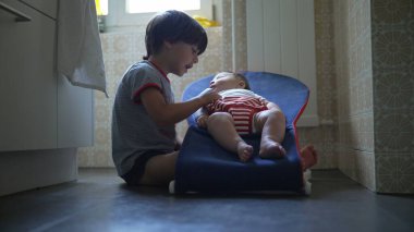 Yerde oturan genç bir çocuk, bir fedainin içinde bebekle konuşuyor, nazikçe iletişim kuruyor ve gülümsüyor, sıcak bir ev ortamında, kardeş bağı kuruyor ve sevgi dolu bir aile ortamında bağ kuruyor.