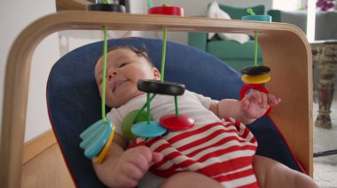 Bebek bir fedaide uzanıyor, renkli asılı oyuncaklarla gülümsüyor ve etkileşimde bulunuyor, merak ve bağlılık gösteriyor, rahat bir ev ortamında, duyusal oyun odaklı.