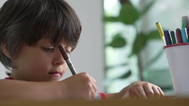 Elinde kahverengi bir kalem tutan çocuk, yanında renkli kalemlerle bir çizim projesine konsantre oluyor.