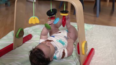 Bebek bir oyun matı üzerinde uzanıyor ve renkli asılı oyuncaklara uzanıyor, sıcak bir aile ortamında oyun saatlerinde erken gelişim ve duyusal keşif ile meşgul oluyor.