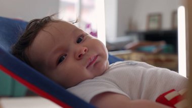 Fedaide uzanmış bebek yakın plan, geniş gözler ve dudaklarla ince yüz ifadeleri yapıyor. Güvenli bir evde sakin ve rahat bir şekilde çevreyi gözlemliyor.
