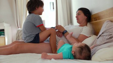 Anne, bebeği ve küçük oğluyla bir yatakta uzanıyor. Rahat bir ev ortamında neşeli bir aile anı yaşıyor.