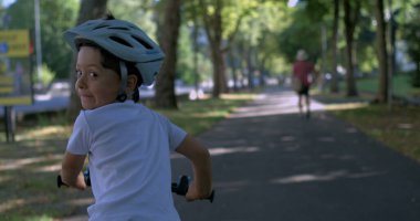 Çocuk kaskla bisiklet sürüyor, gülümsüyor ve arkasına bakıyor, gölgeli park yolunda bir yaz gününün tadını çıkarıyor, 800 fp hızla süper yavaş çekimde, açık hava keyfi ve özgürlüğüyle filme alınıyor.