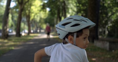 Genç bir çocuk kaskla bisiklete biniyor, neşeli bir ifadeyle geriye bakıyor, yaz boyunca ağaçlık bir yolda, süper yavaş çekim, 800 fps görüntü, açık havada çocukluk eğlencesi ve keşif.