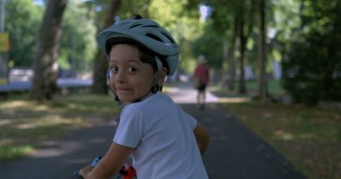 Çocuk kaskla bisiklet sürüyor, gülümsüyor ve arkasına bakıyor, gölgeli park yolunda bir yaz gününün tadını çıkarıyor, 800 fp hızla süper yavaş çekimde, açık hava keyfi ve özgürlüğüyle filme alınıyor.