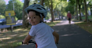 Çocuk kaskla bisiklet sürüyor, gülümsüyor ve arkasına bakıyor, gölgeli park yolunda bir yaz gününün tadını çıkarıyor, 800 fp hızla süper yavaş çekimde, açık hava keyfi ve özgürlüğüyle filme alınıyor.