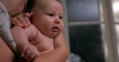 Bebek banyodan sonra tutuluyor, havluya sarılmış, sakin ve tetikte, banyo sonrası bakım için sabırsızlanıyor.