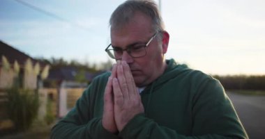 Dışarıda dua eden, elleri sımsıkı bağlı, aşağıya bakan, derin düşüncelere dalmış, günbatımında sakin kırsal bir sahne, minnettarlık ve maneviyat, huzurlu bir atmosfer.