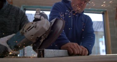 Açı öğütücüyle metal kesen işçiler, başka bir işçi metali sabit tutarken kıvılcımlar uçuşuyor, takım çalışması ve hassasiyeti vurgulanıyor, 800 fps yavaş çekim