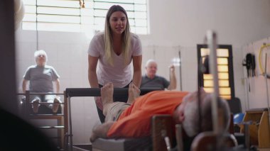 Yaşlı adam pilates makinesinde yattı. Kadın bir pilates koçunun yardımıyla vücudu güçlendirdi. Yaşlılık sporu yaşam tarzı çalışma rutini.
