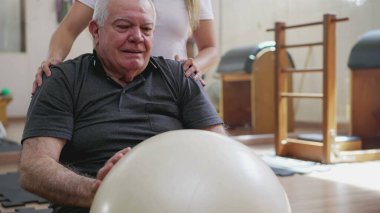 Yaşlı insan, pilates bayan koçunun yardımıyla egzersiz yapıyor. Beyazların kıdemli adamı topla vücudunu güçlendiriyor ve geriyor..