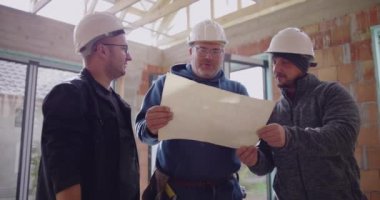 Üç inşaat işçisi gülümseyip bina planlarını tartışırken, inşaat projesiyle ilgili kısmen inşa edilmiş bir yapının planlarını gözden geçiriyorlar.