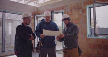 Üç inşaat işçisi planlarını gözden geçiriyorlar, güvenlik kaskı takıyorlar, proje detaylarını tartışıyorlar, takım çalışması ve bitmemiş endüstriyel alanda işbirliği yapıyorlar.