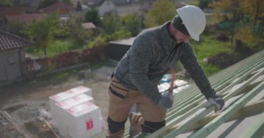 İnşaat işçisi çatıdaki tahta kirişleri çekiç, yan görüş, güvenlik kaskı ve eldiven ile sağlamlaştırıyor yerleşim alanındaki yüksek yapı üzerinde çalışıyor.