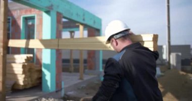 İnşaat işçisi omuzunda uzun ahşap kalaslar taşıyor, inşaat alanı boyunca malzeme taşıyor marangozlukta takım çalışması ve dayanıklılık için.