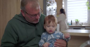 Bebeğin kucağında küçük bir yiyecek tutarken baba sıcak bir mutfak ortamında aile yemeği sırasında samimi ve şefkatli bir an.