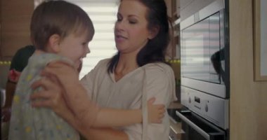 Anne küçük bir çocuğu mutfaktan alıyor, sevgi ve ilgi gösteriyor, arka planda bir adam tezgaha yaslanıyor, sıcak aile anı içeride.