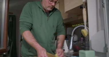 Eller mutfak lavabosunda musluk suyunun altında akan bir süngerle kesme tahtasını ovalayarak modern ev ortamında günlük temizlik işlerini tasvir ediyor.