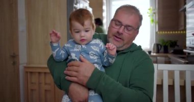 Baba kucağında beşikte, şefkatle aşağıya bakıyor, sıcak bir yuva ortamında şefkatli ve huzurlu bir anı paylaşıyor.