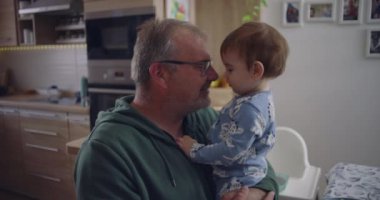 Baba kucağında bebekle, hassas bir anı paylaşıyor, rahat bir ev mutfağında göz teması kuruyor, anne arka planda yemek hazırlıyor.