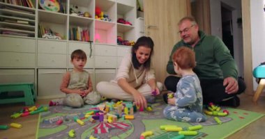 Yerde oturan aile renkli oyuncaklarla ortak blok oyunu oynuyor anne ve baba sıcak ve eğlenceli oturma odasında çocuklarla etkileşime geçiyor.