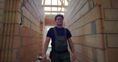 Yeşil tulumlu bir adam bitmemiş binanın koridorunda yürüyor, gözlüklü, odaklanmış bir ifade takınıyor, tuğla duvarları ve ahşap iskeleti var.