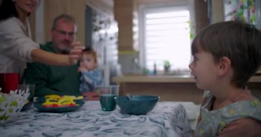 Anne, aile yemeği sırasında gülümseyen çocuğa kurabiye veriyor, baba ve bebekle sıcak bir evde yemek masasında neşeli bir etkileşim.