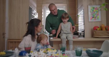 Anne, mutfak tezgahında oturan büyük çocuğa yemek sunarken baba yakınlarda duruyor. Bebek, annesinin kucağında yemek masasındaki aile bağlarını inceliyor. 