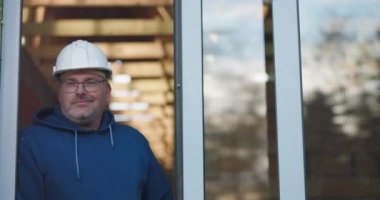 Kapıda duran bir işçi, hafif bir gülümseme, güvenlik kaskı ve gözlük takan, gururlu ama profesyonel bir tavırla inşaat projesini denetleyen