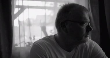 İçerde oturan bir adamın siyah beyaz portresi, pencerenin kenarına dikkatlice bakıyor, perdeler ve yumuşak ışık sakin bir yansıtıcı atmosfer yaratıyor.