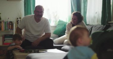 Bebek kanepeye uzanıp aile etkileşimlerini izliyor, baba sehpada çocukla trencilik oynuyor, anne yakınlarda gülümsüyor, aile içi yakınlaşma anı.